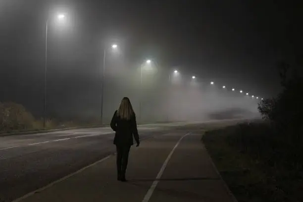 Photo of Young woman alone slowly walking under white street lights in night. Dark time. Peaceful atmosphere in mist. Foggy air. Back view.