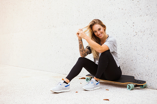 Girl is smiling and sitting on the longboard in front of the wall and enjoying life