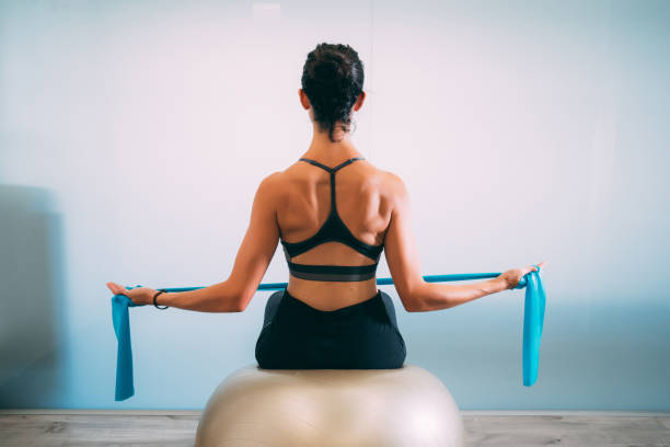 jeune femme séduisante sportive pratiquant le pilates avec les bandes de résistance arrière assis sur les balles de remise en forme - yoga ball photos et images de collection