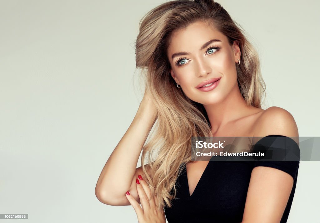 Portrait of gorgeous young woman with elegant make up and perfect golden hairstyle. Young golden haired pretty model tenderly looking aside with light smile and touching  well groomed hair. Portrait of gorgeous young woman with elegant make up and freely lying colored hairstyle. Hair coloration, hair care and cosmetology. Women Stock Photo