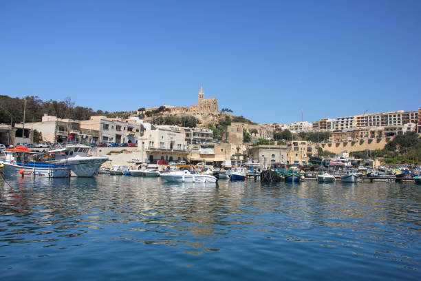 Gozo Ferry Terminal view Mgarr. Bay with yachts on foreground and old city with church on the top on background Mgarr, Malta - May 2018: Gozo Ferry Terminal view Mgarr. Bay with yachts on foreground and old city with church on the top on background. Reflections in blue water mgarr malta island gozo cityscape with harbor stock pictures, royalty-free photos & images