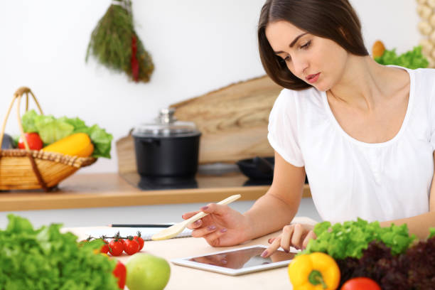 istock Beautiful Hispanic woman cooking in kitchen while using tablet computer and wooden spoon. Housewife found new recipe for dinner or breakfast. Healthy meal and householding concepts 1062473110