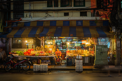 Shopkeeper waiting for customers at his roadside grocery shop illuminated at night in Bangkok’s Chinatown, Thailand.