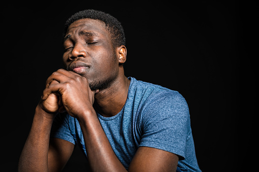 Close-up of thoughtful man with hands clasped. Depressed male is in blue t-shirt. He is against black background.