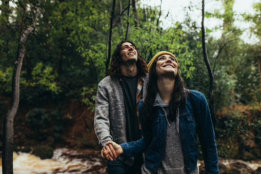 Young woman with her boyfriend in a forest, both looking up and smiling. Happy young couple standing together on rainy day at the forest.