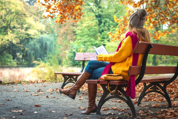 donna seduta in panchina e libro di lettura. autunno al parco pubblico. - autumn women leaf scarf foto e immagini stock