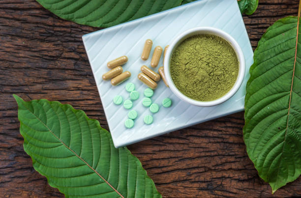 Mitragyna speciosa or kratom leaves with medicinal products in pills, capsules and powder in white ceramic bowl and wooden table, top view stock photo