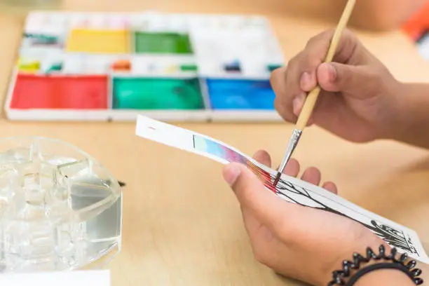 Photo of Arts painting class in school with young student's hand using brush coloring watercolor paint on craft drawing paper