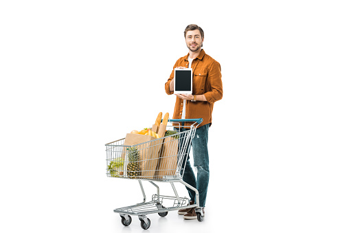 happy man showing digital tablet with blank screen near shopping trolley with paper bags isolated on white