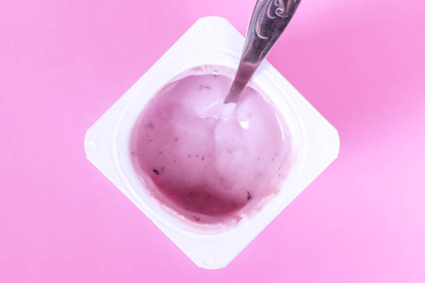 Blueberry yogurt in a plastic cup with a spoon inside on pink background closeup.