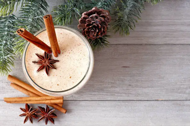 Christmas spiced eggnog in a glass. Top view, with corner border of tree branches on a rustic gray wood background.