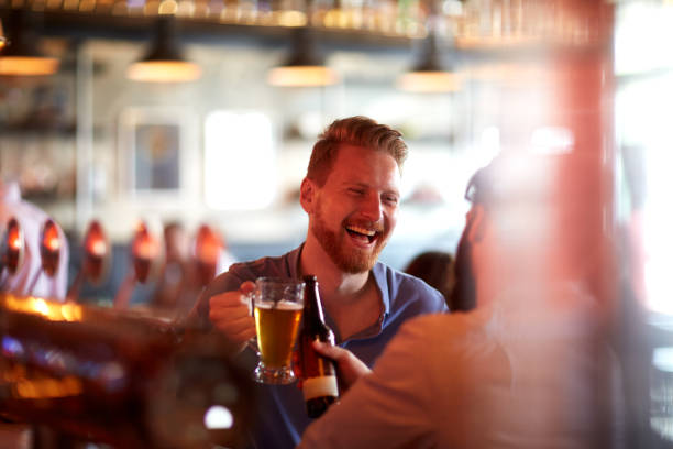 Friends drinking beer at bar counter Bearded male friends standing at the bar counter and having a toast with beer drinks. pub bar counter bar men stock pictures, royalty-free photos & images