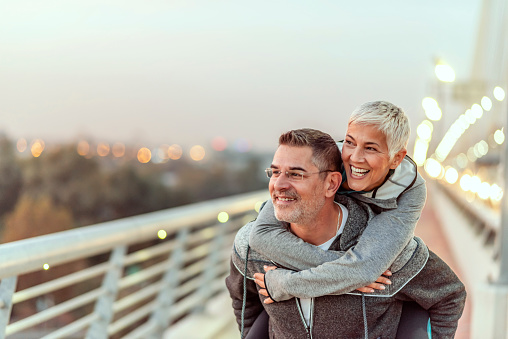 Photo of Happy mature couple having fun, hugging in the city on a autumn day. Love story true feelings concept. Portrait of friendly peaceful fitness couple in love. Couple enjoying the outdoors together. Sporty couple in love enjoying each other. Handsome  man giving a gray hair woman a piggy back ride.