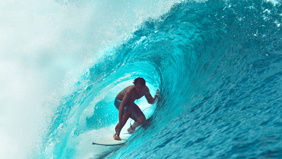 CLOSE UP: Exreme athlete surfs a big barrel ocean wave glistening in the summer sunshine. Spectacular tube wave splashes over the young surfer enjoying his active vacation in sunny French Polynesia.