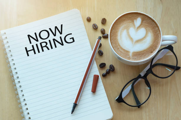istock Concept NOW HIRING message on notebook with glasses, pencil and coffee cup on wooden table. 1063102848