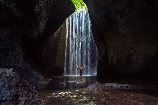 Woman stand in underground cave pool under falling fresh water of Tukad Cepung waterfall. Nature day tour, hiking activity adventure and fun at family tourist camp on summer vacation in Bali island