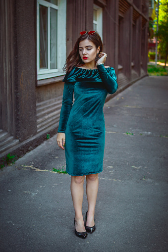 Portrait of beautiful young brunette woman with bright makeup, wearing emerald green velvet dress standing near old brown building