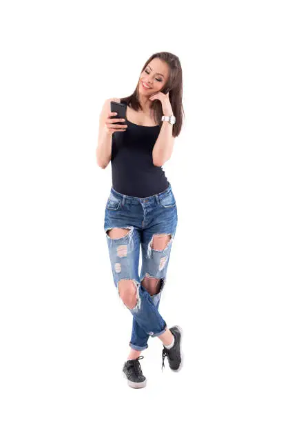 Trendy stylish modern pretty young woman reading message on cellphone in ripped jeans. Full body isolated on white background.