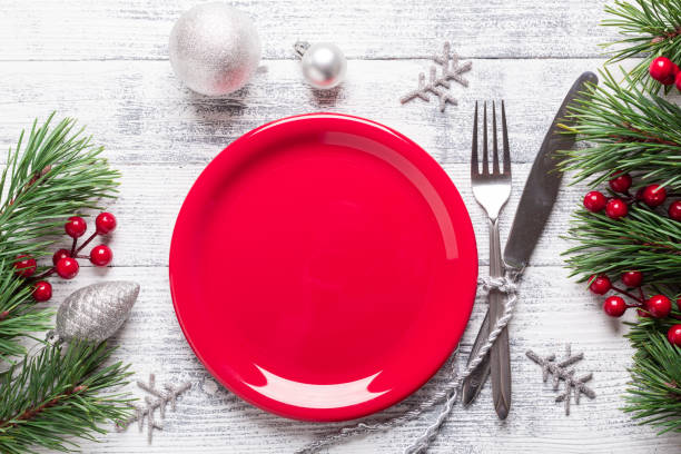 istock Christmas table setting with empty red plate, gift box and silverware on light wood background. Fir tree branch, holly berries. 1063235284