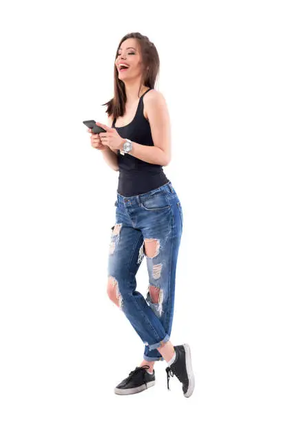 Pretty modern young woman laughing heartily white holding cellphone in trendy torn jeans. Full body isolated on white background.