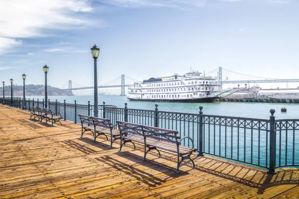 Benches at historic Pier 7 with traditional paddleboat and Oakland Bay Bridge in the background on a sunny day with bliue sky and clouds, San Francisco, California, USA
