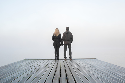 Man and woman holding each other hands. Young couple standing on edge of footbridge. Foggy air. Early chilly morning in autumn. Peaceful atmosphere in mist. Back view.