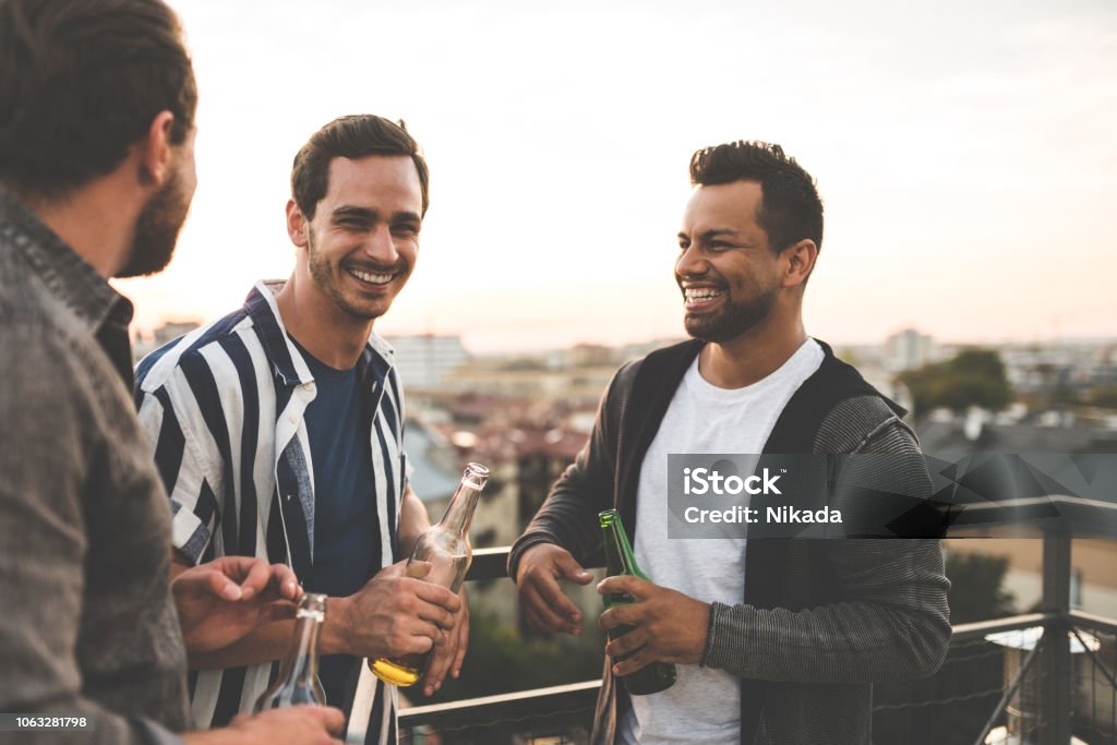 Cheerful friends enjoying at rooftop party Group of male friends standing together at rooftop with drinks and chatting. Cheerful friends enjoying at terrace party. Friendship Stock Photo