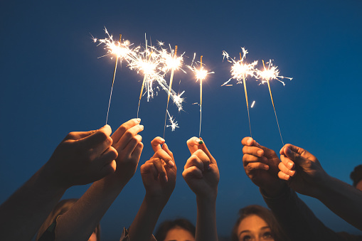 Close up of young people hands holding sparklers against sky. Friends celebrating new years eve with fireworks.