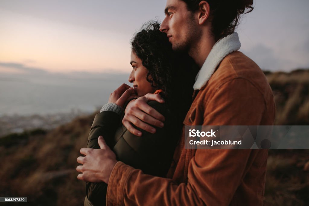 Romantic young couple standing in mountain Romantic young couple standing in mountain together and looking at view. Young man embracing his girlfriend and looking away. Love - Emotion Stock Photo