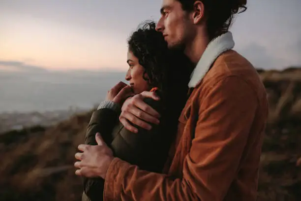 Romantic young couple standing in mountain together and looking at view. Young man embracing his girlfriend and looking away.