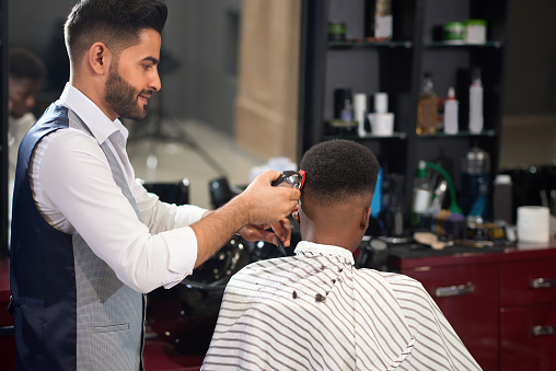Stylish Hairdresser In White Shirt And Waistcoat Trimming Hair Of Male Client With Clipper In Barber Shop Man Sitting On Chair And Getting Trendy Haircut Concept Of Hairstyle And Shave Stock Photo - Stylish Hairdresser In White Shirt And Waistcoat Trimming Hair Of Male Client With Clipper In Barber Shop Man Sitting On Chair And Getting Trendy Haircut Concept Of Hairstyle And Shave Stock Photo -