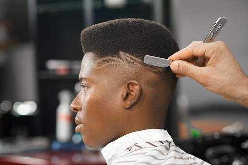 Side view of serious man with stylish modern haircut looking forward in barber shop. Hand of barber keeping straight razor and cutting trendy stripes on head of client. Concept of shaving.