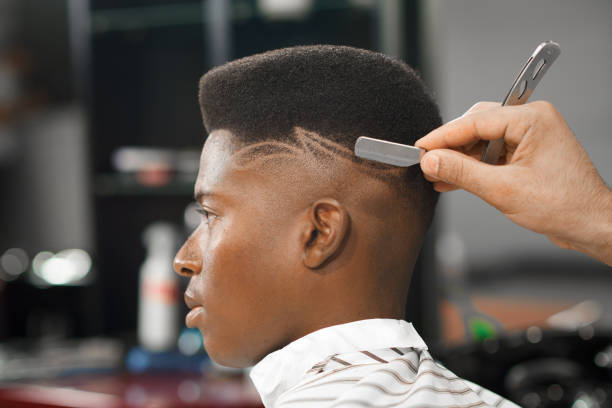 Side view of serious man with stylish modern haircut looking forward in barber shop. Hand of barber keeping straight razor and cutting trendy stripes on head of client. Concept of shaving. stock photo