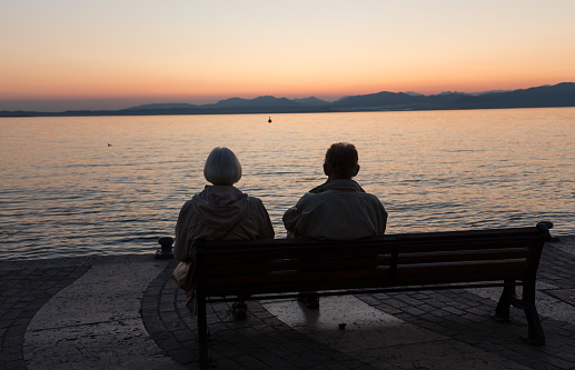 Aged couple sitting on a bench and looking at sunset at Garda Lake, Italy