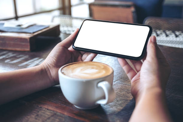 istock Mockup image of hands holding and using a black mobile phone with blank screen horizontally for watching with coffee cup on wooden table 1063318526