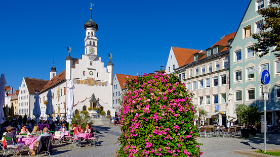 People sitting at the outdoors tables in the Market Square in Kempten, the oldest urban settlement in Germany. On the square overlooks the town hall, a medieval building built in 1474, with the current facade dating to the 1930s.