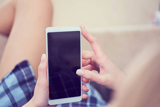 istock Back view of a woman hands using smart phone with blank screen close up. White phone with a black screen in the hands of a young girl lying on the floor with her feet on the couch 1063342550