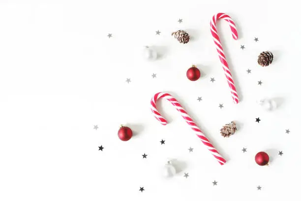 Christmas styled composition. Christmas red and white balls, baubles, candy canes, glittering silver stars confetti decoration and larch cones on white background, flat lay, top view. Winter patttern.