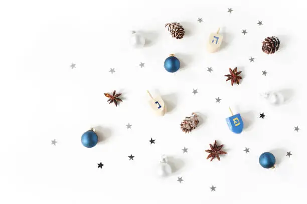 Hanukkah styled stock composition. Decorative pattern. Wooden dreidel toys, larch cones, anise and silver confetti stars decoration on white background, flat lay, top view. Jewish design.