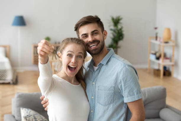 Couple in love standing in living room at new house Smiling married couple in love standing hugging in living room at new modern house looking at camera. Excited wife holding keys from their new dwelling. Happy family, moving real estate owners concept happy family with key of their new house stock pictures, royalty-free photos & images