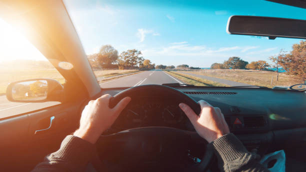 Point of view seen from driver holding on to steering wheel of a car Personal perspective - point of view (POV) - of driver, who drives his car on a road. The hands are on the steering wheel and the sun is shining outside. He drives on country roads as the sun is about to set. The sky is blue. personal perspective stock pictures, royalty-free photos & images