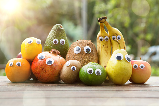 Funny fruit and vegetables with eyes .Orange, lemon, green apple, red apple, avocado , banana, pomegranate , coconut, kiwi, tangerine , pear on wooden table in public park.Cute food with faces.Funny fruit characters.