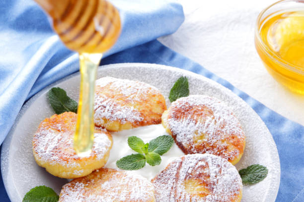 istock Cottage cheese pancakes sprinkled powdered sugar and fresh mint. Cheesecakes on white plate. Sour cream in wooden spoon. Honey and dairy products on white blue background. Healthy food 1063475184