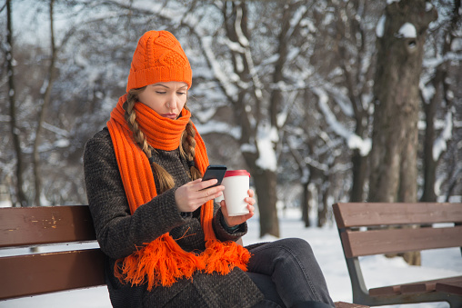 A young woman with braided long blond hair and a nostril piercing is in a outdoors park during the winter, sitting on a bench. There is snow and trees in the background. She is wearing winter clothes, an orange hat and scarf, and is holding a disposable cup with a hot drink. She is using a smartphone. With copy space.