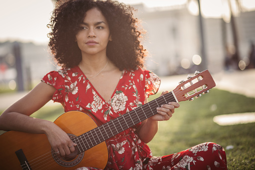 One woman, young and beautiful, sitting in park, playing acoustic guitar.