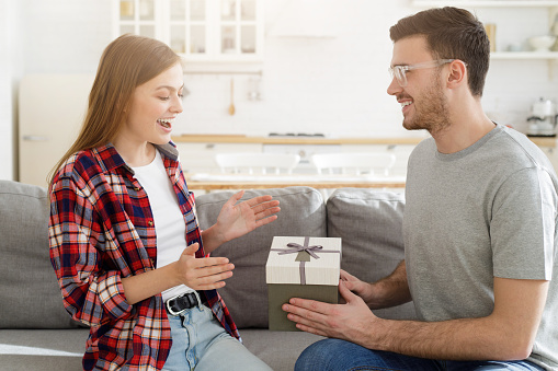 Handsome young man is giving gift box to surprised girlfriend at home