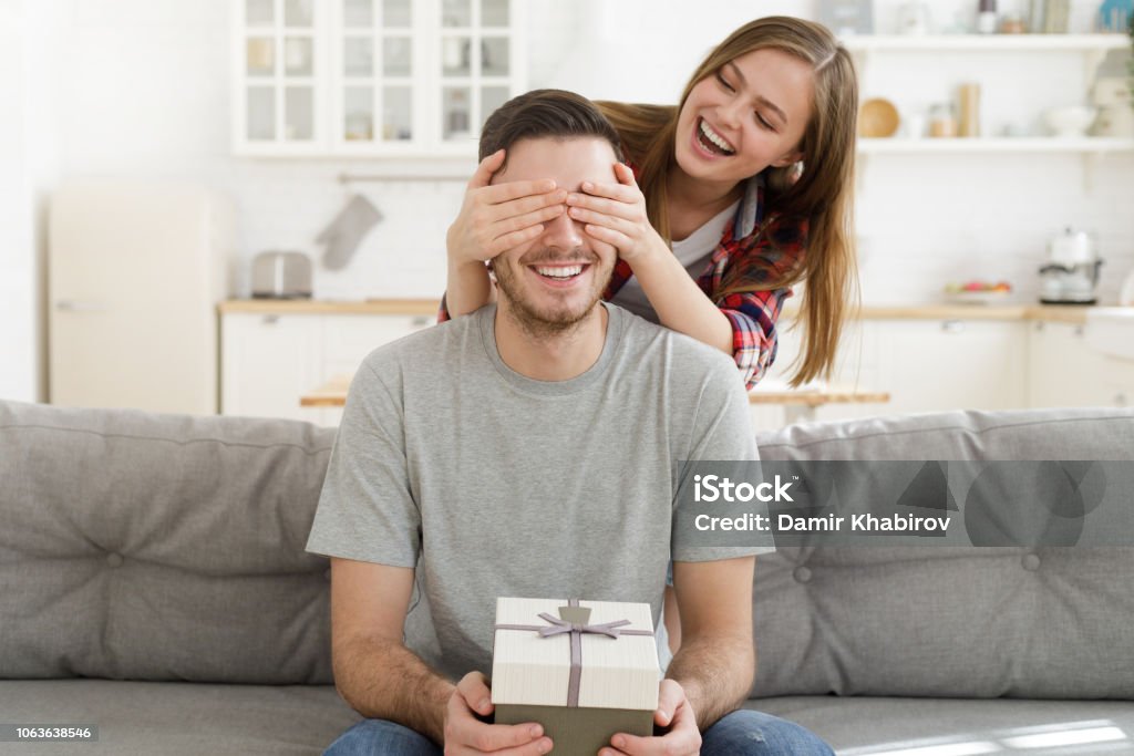 Giovane donna tiene gli occhi chiusi al suo ragazzo a casa, mentre è seduto sul divano con una confezione regalo, festeggiando il compleanno - Foto stock royalty-free di Regalo