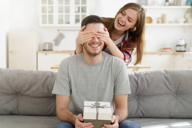 Photo of Young female keeps eyes closed to his boyfriend at home, while he is sitting on sofa with gift box, celebrating birthday