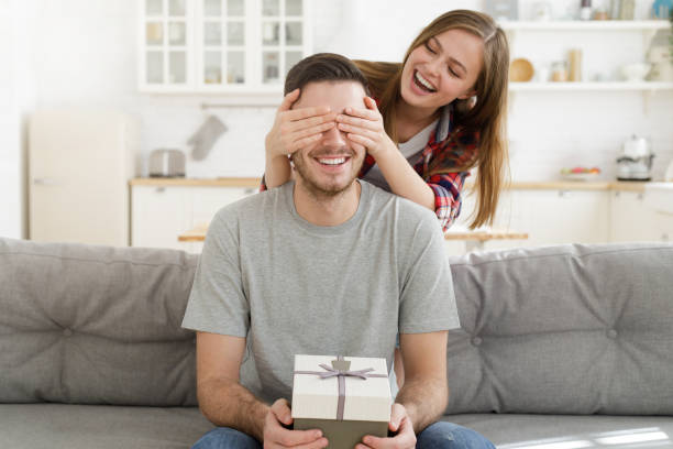 young female keeps eyes closed to his boyfriend at home, while he is sitting on sofa with gift box, celebrating birthday - anniversary present imagens e fotografias de stock