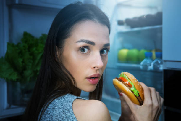 donna affamata che mangia hamburger di notte vicino al frigorifero - ingordigia foto e immagini stock